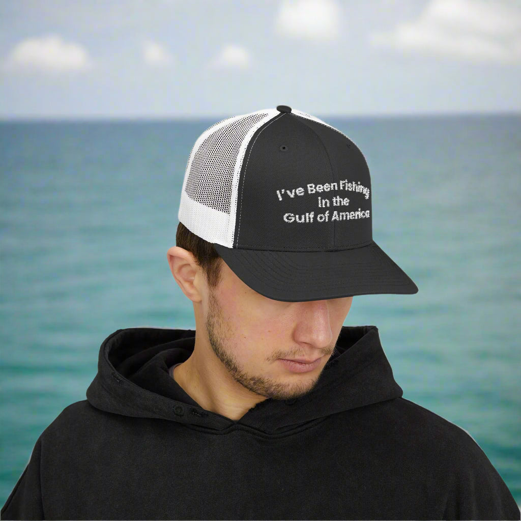 Snapback trucker cap with “I’ve Been Fishing the Gulf of America” embroidered on the front, resting on a boat deck with Gulf waters in the background
