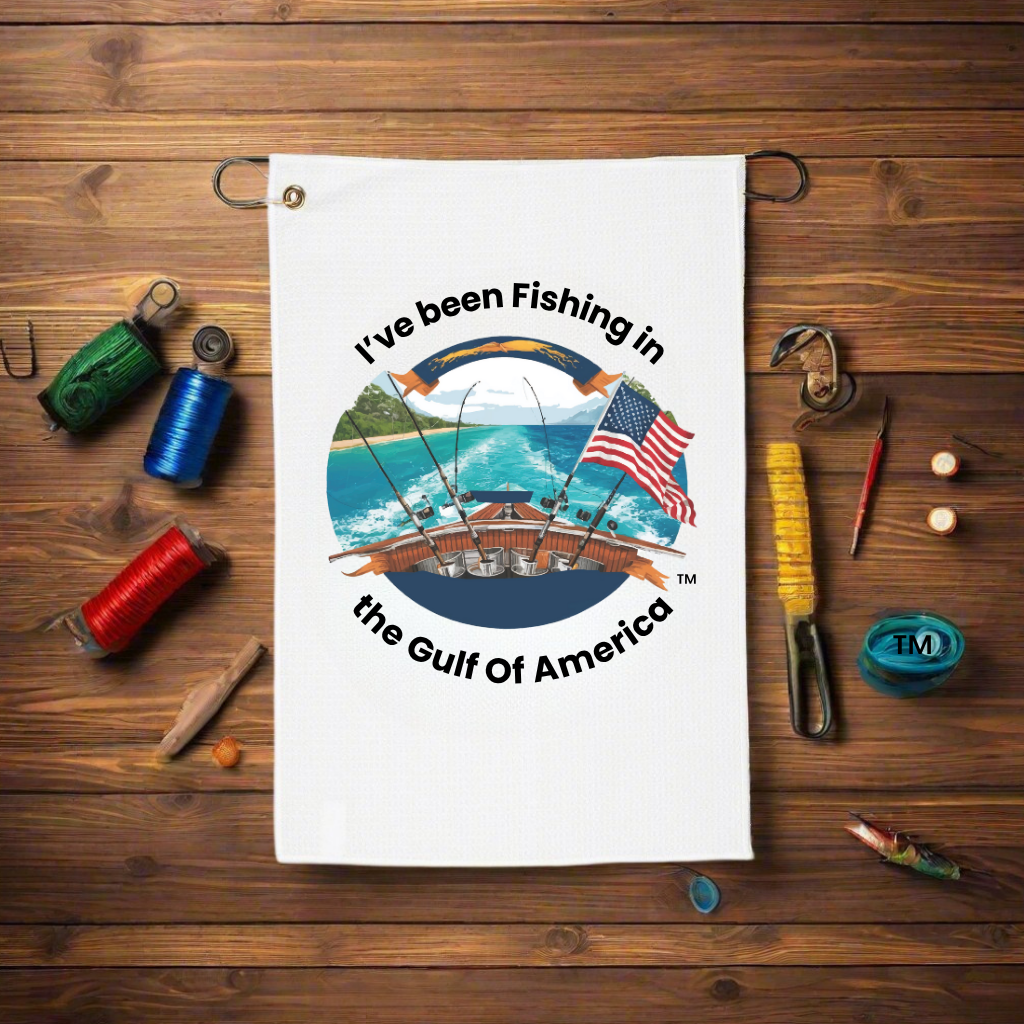 Golf towel with “I’ve Been Fishing in the Gulf of America” text, clipped to a golf bag on a course with coastal scenery in the background